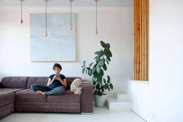 Young woman with short hair style siting on couch and talking on phone at home. Housewife relaxing and chatting through mobile phone. Interior design. Copy space.