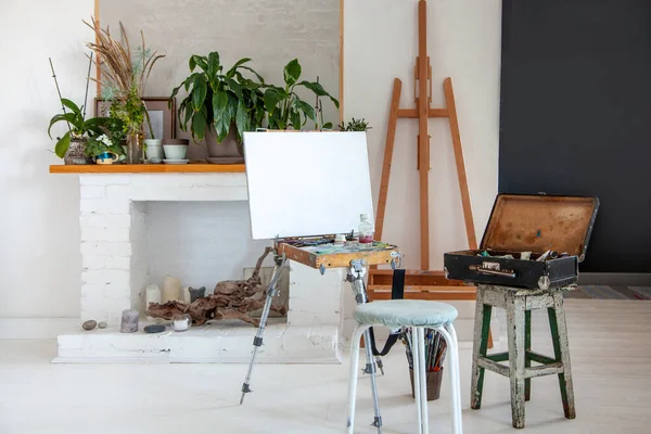 Art studio interior design with painting supplies, canvas on easel, paintbrushes, vintage oil paint box, palette near the decorative fireplace and potted plants. Painter tools in workshop. Copy space