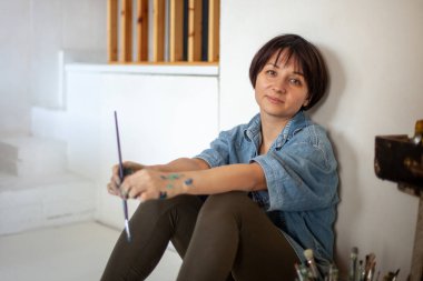 Boya fırçası ile oturan kadın ressam, boya fırçalı kova ve aletlerle birlikte eski tip ahşap kutunun yanındaki sehpada resim yapmaya çalışıyor. Ressam onun sanat eserlerini stüdyoda analiz ediyor.
