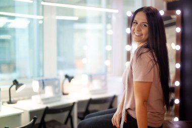 Şablon masasında oturan mutlu genç kadın atölyedeki dikiş makinelerinin yanında kumaş kesiyor. Genç terzi ustalık kursundan sonra dinleniyor. Büyük pencereli moda atölyesi iç mimarisi.