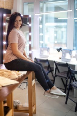 Şablon masasında oturan mutlu genç kadın atölyedeki dikiş makinelerinin yanında kumaş kesiyor. Genç terzi ustalık kursundan sonra dinleniyor. Büyük pencereli moda atölyesi iç mimarisi.