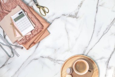 Katlanmış mercan rengi dantel ve iplik makaralı ipek kumaşlar, hesap makinesi, not defteri, kalem, mezura, kahve fincanının yanında altın tepside kurabiyeler olan makas. Boşluğu kopyala
