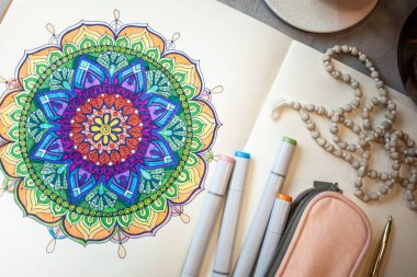 Renkli mandala sanat kitabının üst görüntüsü renkli kalemler, pembe kalem kutusu ve tespih boncukları. Meditasyon tekniği olarak renkli mandalalar kavramı.