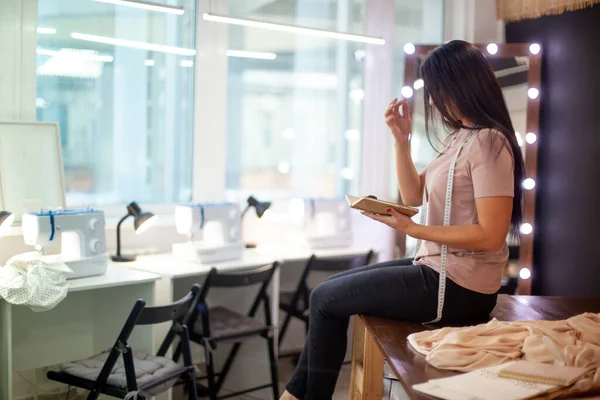 Şablon masasında oturan mutlu genç kadın atölyedeki dikiş makinelerinin yanında kumaş kesiyor. Genç terzi ustalık kursundan sonra dinleniyor. Büyük pencereli moda atölyesi iç mimarisi.