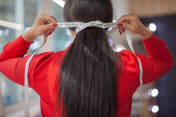 Female fashion designer in red dress wrapped her dark long hair with tape measure. Well groomed woman tailor hands measuring the thickness of the hair. Beauty and haircare concept