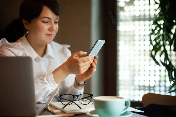 コーヒーを飲みながら携帯電話からのテキストメッセージを読んでビジネスホワイトブラウスの若い女性 ビジネスマンは彼女のスマートフォンでソーシャルメディアをチェックしています ブロガーのライフスタイル 計画と組織 — ストック写真