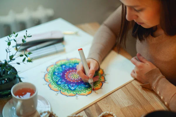 Young woman colouring mandala with markers and white rosary on table with cup of coffee near flower vase at home. Art therapy and meditation concept. Hobby and leisure during quarantine. Wellbeing.