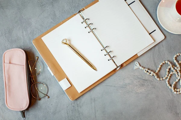 Workspace with opened notebook with clean sheets, gold pen, rosary beads, pink colored case and cup of tea near vase with decorative plants. Religions, culture and education concept. Copy space.