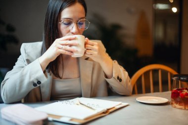 Koyu renk takım elbiseli, genç ve güzel bir kadın tatil zamanında kahve içerken iş planlarını günlüğe yazıyor. Planlama ve zaman yönetimi konsepti. Olumlu bir tavırla öğrenmek.