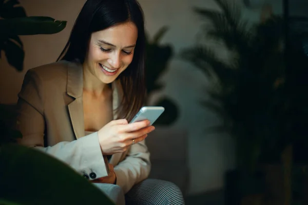 穿着西服的漂亮女商人正坐在沙发上 微笑着在手机上阅读和输入短信 销售通信和信息概念 企业家 自由撰稿人 复制空间 — 图库照片