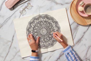 Renkli mandala sanat kitabının en üst görüntüsü. Renkli kalemler, pembe kalem kutusu, bir fincan kahve ve kurabiye. Beyaz mermer arka plan. Kız çiziyor. Eller yukarı.