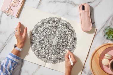 Renkli mandala sanat kitabının en üst görüntüsü. Renkli kalemler, pembe kalem kutusu, bir fincan kahve ve kurabiye. Beyaz mermer arka plan. Kız çiziyor. Eller yukarı.