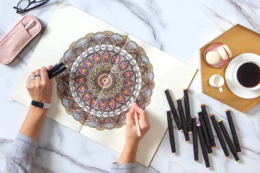 Kadın eskiz defterinde renkli kalemlerle mandala çiziyor ve boyuyor. Kahvaltlarını kahve ve mermer masada badem kurabiyeleriyle yapıyor. Resim yaparak stresi azaltma eğilimi. Sanat terapisi. Boş zaman.