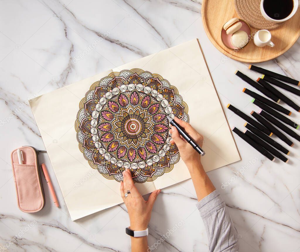 Woman drawing and coloring mandala in sketchbook with colorful markers while having breakfast with coffee and macarons on white marble table. Stress relieving trend by painting. Art therapy. Leisure.