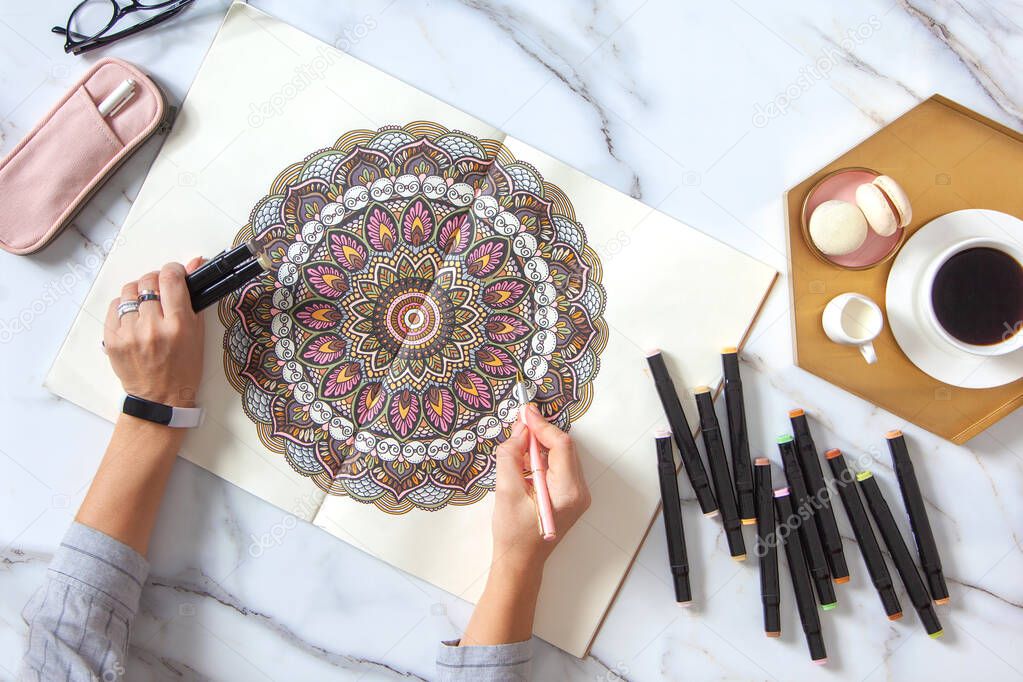 Woman drawing and coloring mandala in sketchbook with colorful markers while having breakfast with coffee and macarons on white marble table. Stress relieving trend by painting. Art therapy. Leisure.
