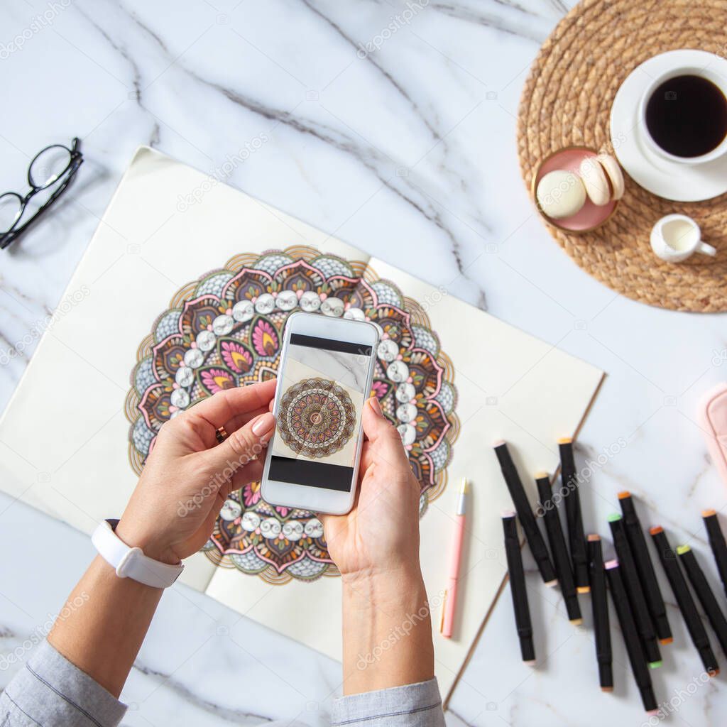 Woman is taking photos of colored mandala from sketchbook on white marble desk with displayed markers, eyeglasses and cup of coffee. Social media. Meditation. Ethnic oriental circle ornament.