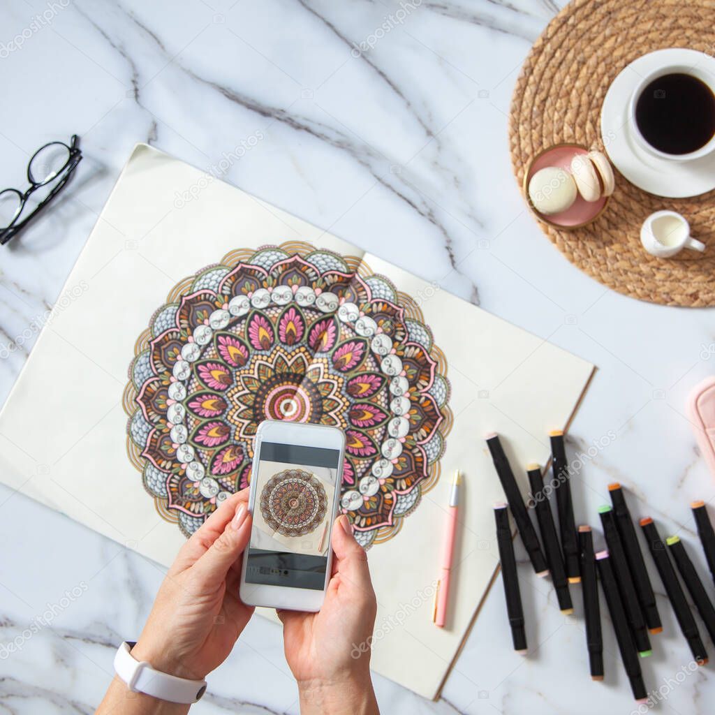 Woman is taking photos of colored mandala from sketchbook on white marble desk with displayed markers, eyeglasses and cup of coffee. Social media. Meditation. Ethnic oriental circle ornament.