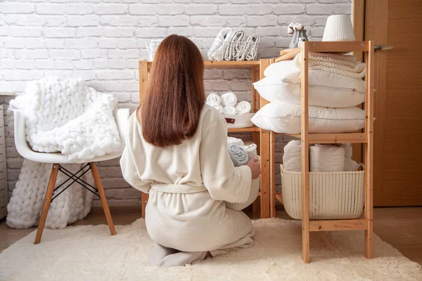 Young woman in warm robe is organizing neatly folded towels, sheets, blankets in white wicker and steel wire baskets and placing on wooden shelves. Konmari method of linen closet organization concept.