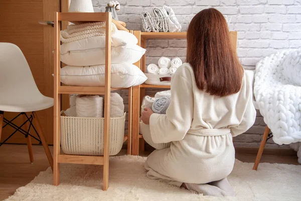 Young woman in warm robe is organizing neatly folded towels, sheets, blankets in white wicker and steel wire baskets and placing on wooden shelves. Konmari method of linen closet organization concept.