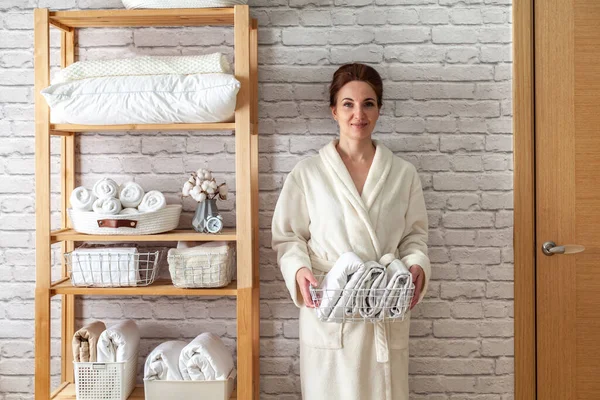 stock image Woman in robe is standing and holding white wire basket of folded bed sheets near organized linen closet in bathroom. Sorted and folded towels in white wicker and wire baskets placed on wooden shelves