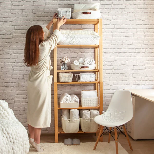 Young woman in warm robe is organizing neatly folded towels, sheets, blankets in white wicker and steel wire baskets and placing on wooden shelves. Konmari method of linen closet organization concept.