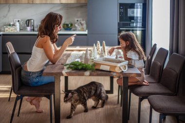Küçük kız Noel Ağacı konisi yapıyor ve annesi mutfak masasının altında yürürken telefonda yazı yazarken düğme ve iplikle süsleme yapıyor. Yeni yıl ve Noel partisi hazırlığı