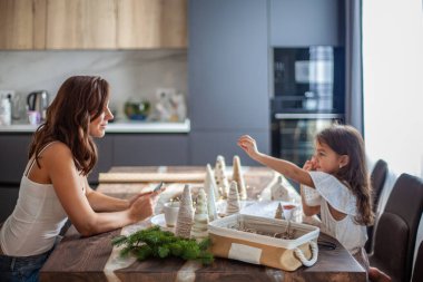 Küçük kız Noel Ağacı konisi yapıyor ve annesi mutfak masasının altında yürürken telefonda yazı yazarken düğme ve iplikle süsleme yapıyor. Yeni yıl ve Noel partisi hazırlığı