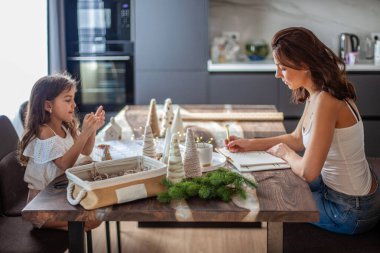 Küçük kız Noel Ağacı konisi yapıyor ve annesi mutfak masasının altında yürürken telefonda yazı yazarken düğme ve iplikle süsleme yapıyor. Yeni yıl ve Noel partisi hazırlığı