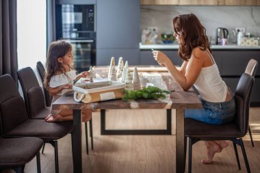 Küçük kız Noel Ağacı konisi yapıyor ve annesi mutfak masasının altında yürürken telefonda yazı yazarken düğme ve iplikle süsleme yapıyor. Yeni yıl ve Noel partisi hazırlığı