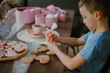 Küçük tatlı çocuk, pembe şeker kremalı zencefilli kek kalbini dekore ediyor. Sevgililer Günü için hazırlık konsepti. Tatlı kornet krema şekeri yer.