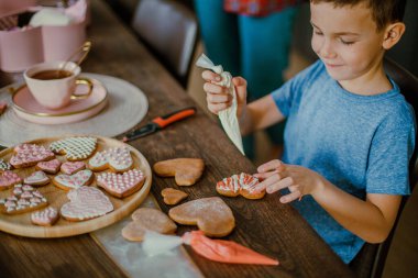 Küçük tatlı çocuk, pembe şeker kremalı zencefilli kek kalbini dekore ediyor. Sevgililer Günü için hazırlık konsepti. Tatlı kornet krema şekeri yer.
