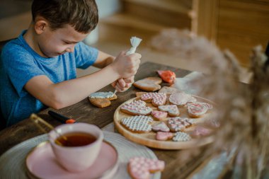 Küçük tatlı çocuk, pembe şeker kremalı zencefilli kek kalbini dekore ediyor. Sevgililer Günü için hazırlık konsepti. Tatlı kornet krema şekeri yer.