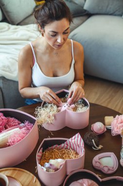 Genç, güzel bir kadın beyaz mermer arka planda kadınlar için pembe renkli kalp şeklinde hediye kutuları, ev yapımı banyo malzemeleri, lokum ve vücut bakım ürünleri açıyor. Sevgililer Günü hediyesi kavramı.