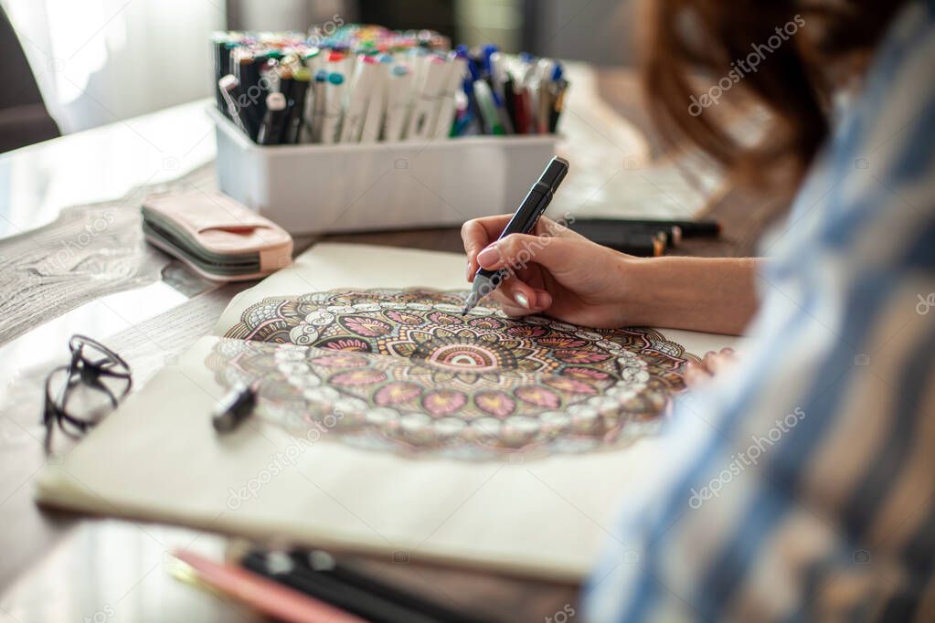 Beautiful well-groomed young woman draws a circular mandala pattern in the album. Artistic markers and colored pencils. Hobby, therapy and home recreation concept.