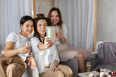 Üç bayan arkadaştan oluşan bir grup ev partisinde fotoğraf çektirip akıllı telefon kullanıyor. Cep telefonuyla selfie çeken mutlu kadınlar evde şampanya içerek eğleniyorlar.