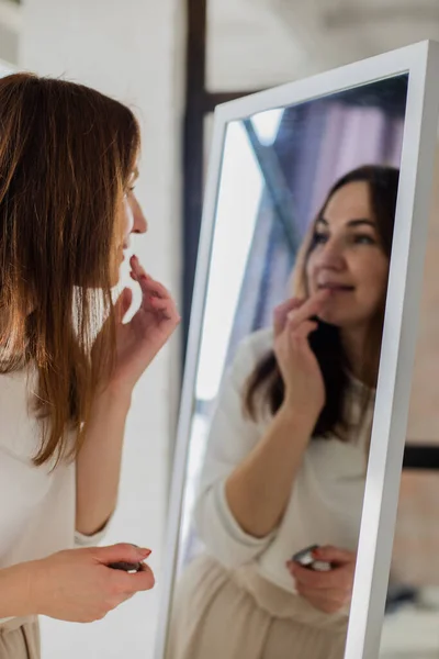 Cheerful brunette woman applying lipstick balm on lips in front of mirror. Beautiful female use dyeing cosmetics or moisturising skin. Pretty lady finishing makeup enjoying facial beauty care