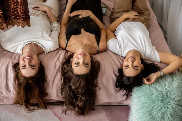 Üç mutlu kadın baş aşağı yatıp gülümsüyor birlikte rahatlıyor ve ev partisinde olumlu duygular yaşıyor. Neşeli, uzun saçlı, rahat yatak odasında poz veren yüksek açılı bir bayan arkadaş.