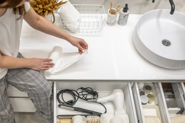 Woman rolling up hand towel and neatly putting into drawer together with toiletries by sitting on bathroom drawer. Concept of organization of bath amenities in storage cabinet under the bathroom sink.