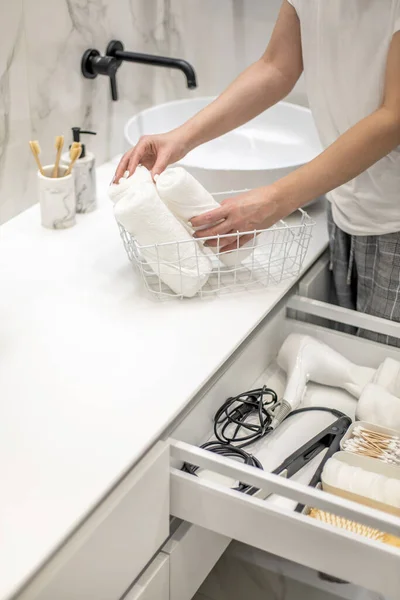 Woman rolling up hand towel and neatly putting into drawer together with toiletries by sitting on bathroom drawer. Concept of organization of bath amenities in storage cabinet under the bathroom sink.