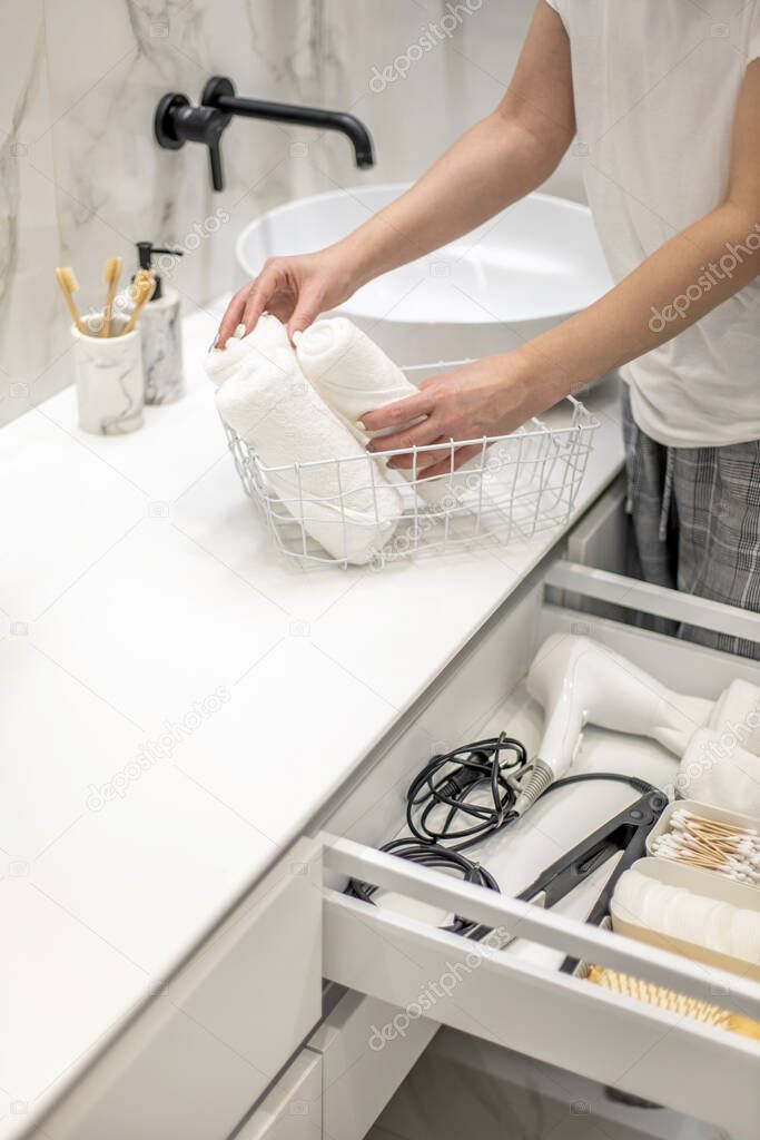 Woman rolling up hand towel and neatly putting into drawer together with toiletries by sitting on bathroom drawer. Concept of organization of bath amenities in storage cabinet under the bathroom sink.