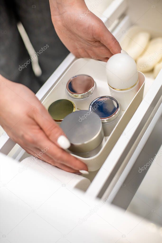 Woman hands holding plastic container with creams and placing into the bathroom drawer. Concept of using toiletry organizer for keeping or organizing personal hygiene accessories with KonMari method.