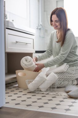 Gülümseyen genç ev hanımı banyo havlusunu sardı ve lavabonun altındaki dolabı düzenledi. Ev işlerinden hoşlanan mutlu bir kadın hijyen pamuğu aksesuarlarını klozete koyuyor. Modern Marie Kondo 'nun yöntemi