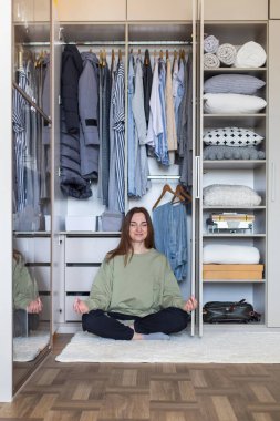 Modern gardırop deposunda nilüfer çiçeği pozisyonunda neşeli bir kadın. Mutlu ev hanımı kıyafetlerle dolu dolapta meditasyon yapıyor. Gülümseyen kadın ev temizliğinden sonra dinleniyor.