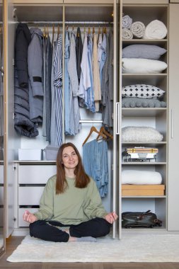 Modern gardırop deposunda nilüfer çiçeği pozisyonunda neşeli bir kadın. Mutlu ev hanımı kıyafetlerle dolu dolapta meditasyon yapıyor. Gülümseyen kadın ev temizliğinden sonra dinleniyor.