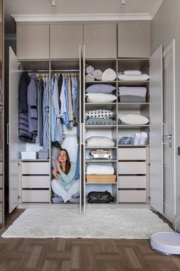 Pijamalı gülümseyen ev hanımı gardıropta yastıkta uyuyor. Modern dolabın içinde kıyafetlerle saklanan eğlenceli, genç bir kadın. Memnuniyet verici ev hanımı hafta sonu evde eğleniyor.