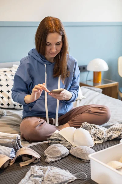 Young woman organizing underwear storage sitting on bed at comfortable cozy bedroom. Domestic casual female putting clothes into boxes wardrobe. Contemporary folded container cases for clothing