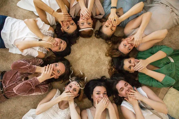 Top view playful female friends hiding mouth by hands laughing relaxing lying circle together at hen-party. Group of funny woman in hippie apparel covering lips face having fun and positive emotion