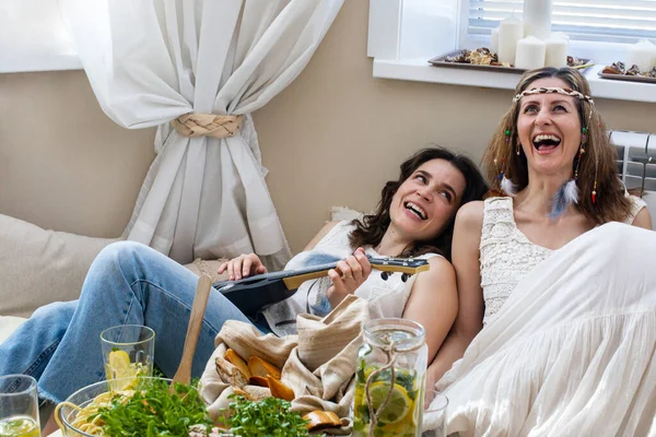 Duas amigas hippies relaxantes cantando música tocando guitarra deitada em almofadas confortáveis servindo mesa — Fotografia de Stock