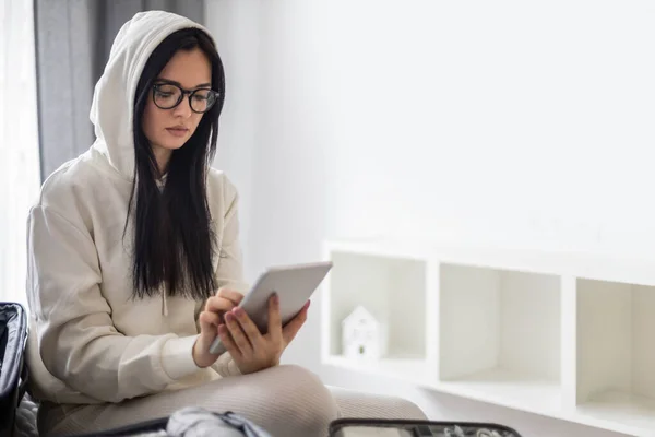 A beautiful young girl in a white casual suit sits on the bed and uses a tablet. — 图库照片