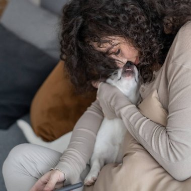 Neşeli kıvırcık kadın öpüyor küçük köpeğe sarılıyor sıcak, minimalist evde sevgi ve şefkat hissediyor.